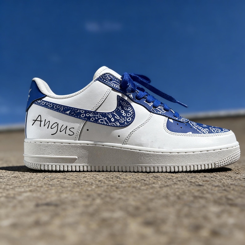 May include: White and blue custom sneaker with a paisley design. The shoe features blue laces and accents, with the name "Angus" written in black on the side. The sneaker is set against a bright blue sky.