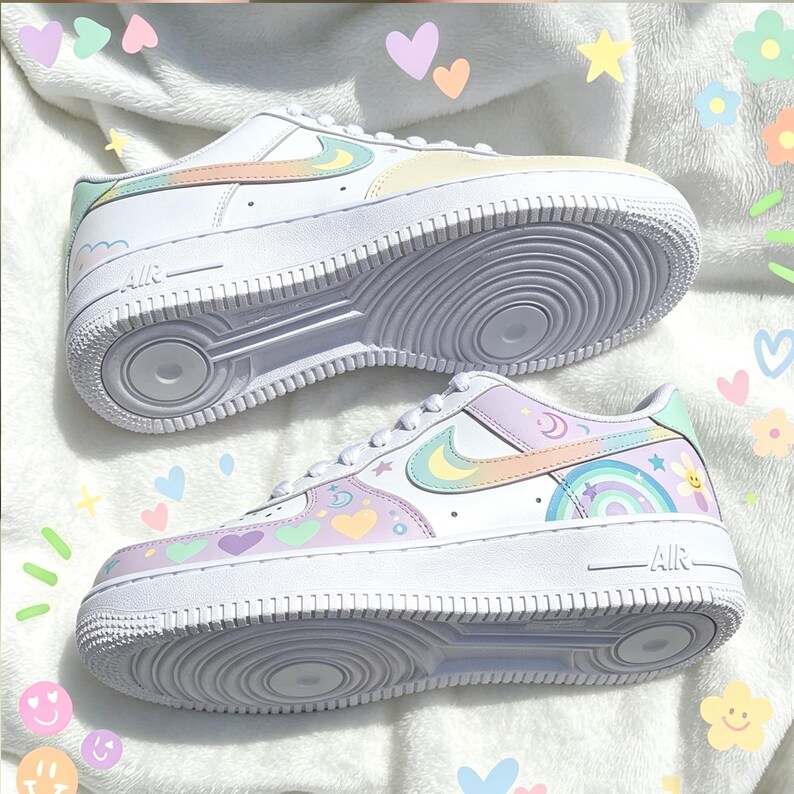 May include: Pair of white sneakers with pastel rainbow and heart designs. The shoes feature a white sole and a colorful swoosh logo. The design includes a rainbow, stars, and crescent moons.