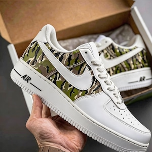 May include: White sneakers with a camouflage pattern on the side panels. The shoes feature a white sole, laces, and a large white Nike swoosh. The word "AIR" is printed in black on the side.