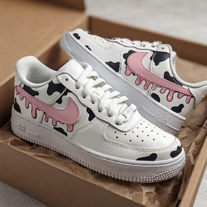 Op de afbeelding: Witte sneakers met zwarte koeienvlekken en roze swooshes. De schoenen hebben een uniek ontwerp met een druipend roze verfeffect langs de zijkanten. De sneakers liggen in een bruine doos.