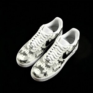 May include: Pair of white sneakers with a camouflage pattern in shades of gray and black. The shoes feature white laces, a black swoosh logo, and a white sole. The sneakers are set against a black background.