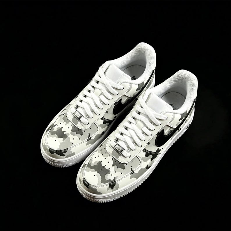 May include: Pair of white sneakers with a camouflage pattern in shades of gray and black. The shoes feature white laces, a black swoosh logo, and a white sole. The sneakers are set against a black background.