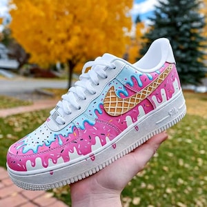 May include: White and pink Air Force 1 sneakers with a unique ice cream design. The shoes feature a white base with pink and blue dripping paint effects, a waffle cone Nike logo, and colorful sprinkles.