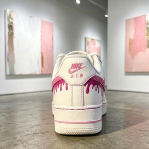 May include: White Nike Air Force 1 sneakers with pink accents. The back of the shoe features the Nike logo and the word "AIR" in pink. Pink paint drip design on the back and side of the shoe.