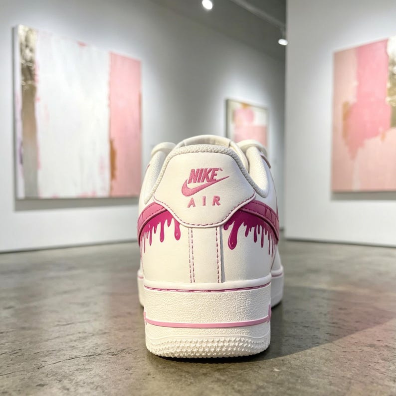 May include: White Nike Air Force 1 sneakers with pink accents. The back of the shoe features the Nike logo and the word "AIR" in pink. Pink paint drip design on the back and side of the shoe.