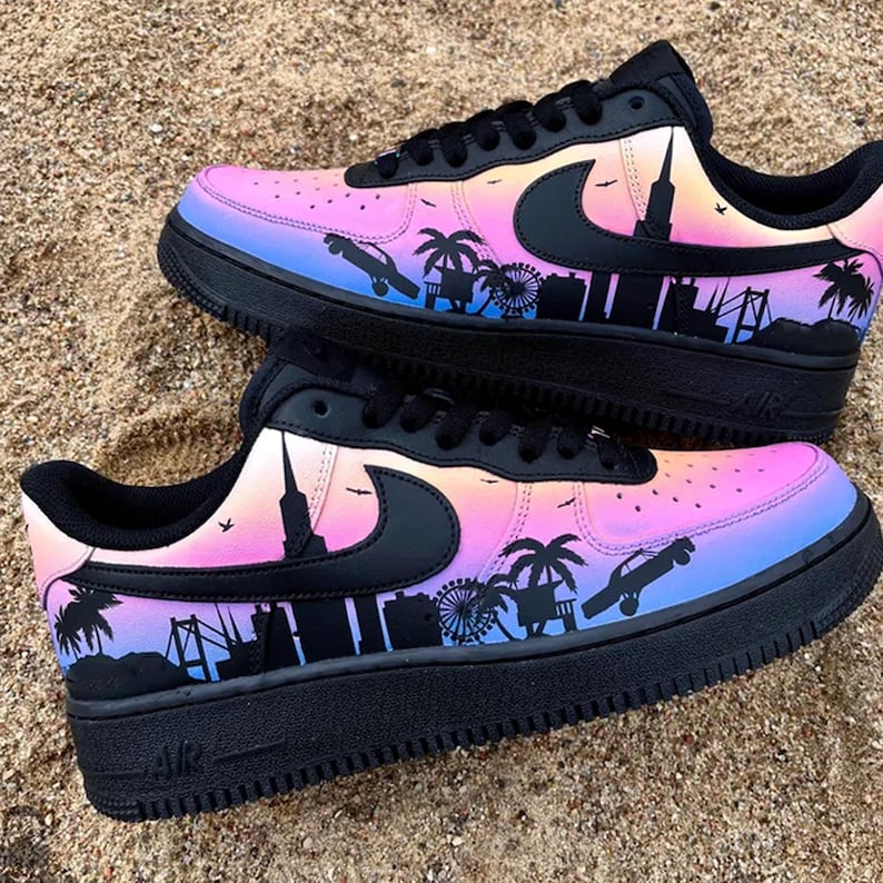 May include: Pair of black sneakers with a colorful gradient design of pink, purple, and blue. The shoes feature a black cityscape silhouette with palm trees, a Ferris wheel, and a car. The Nike swoosh is black.