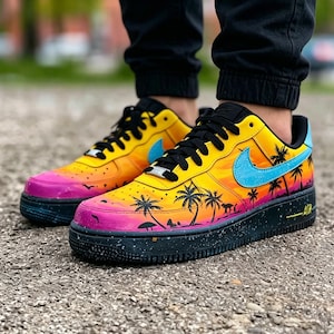May include: Customized sneakers with a vibrant sunset design. The shoes feature a gradient of yellow, orange, and pink, with black palm tree silhouettes. A light blue swoosh and black laces complete the look. The black sole has white speckles.