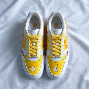 May include: Pair of white sneakers with yellow accents and cartoon character illustrations. The toe box and heel feature yellow designs, while the laces are yellow. The sneakers have a white sole and a cartoon character on the tongue.