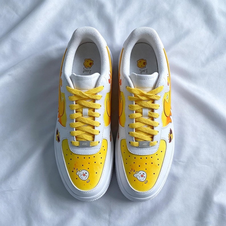 May include: Pair of white sneakers with yellow accents and cartoon character illustrations. The toe box and heel feature yellow designs, while the laces are yellow. The sneakers have a white sole and a cartoon character on the tongue.