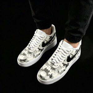 May include: Pair of white and gray camouflage sneakers with black swoosh logos. The shoes have white laces and soles. The sneakers are a classic athletic style.