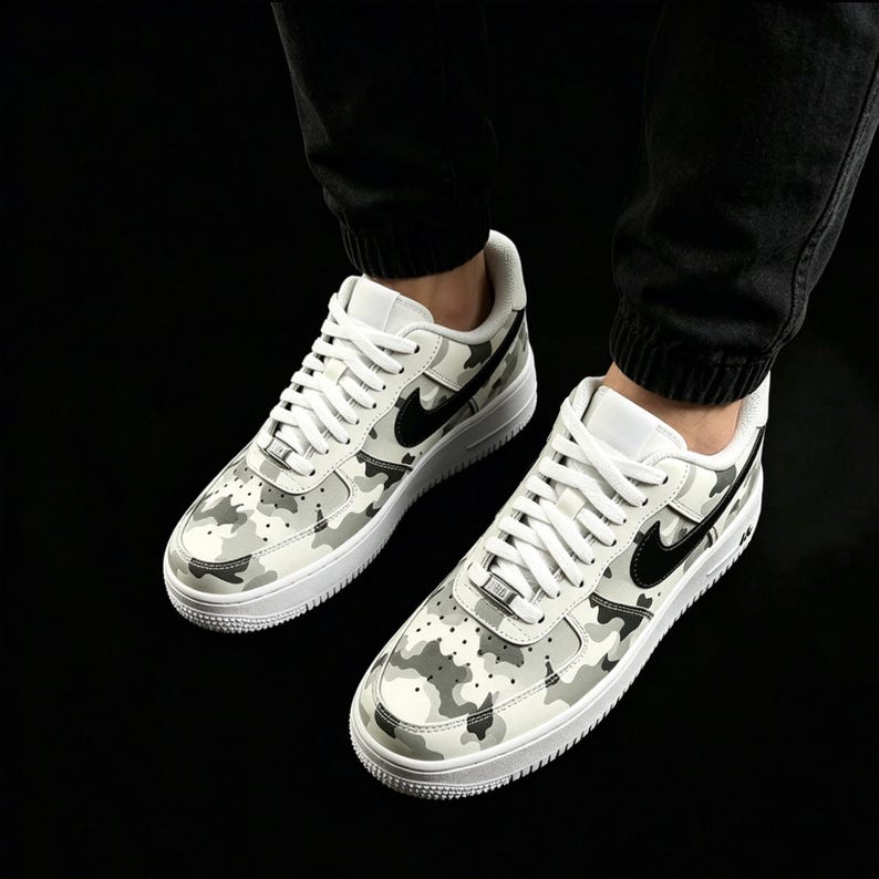 May include: Pair of white and gray camouflage sneakers with black swoosh logos. The shoes have white laces and soles. The sneakers are a classic athletic style.