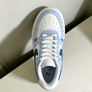 May include: A white and light blue sneaker with a unique design. The shoe features a white base with light blue accents on the toe, sides, and heel. The design includes small blue and green dots and a blue swoosh logo. White laces complete the look.