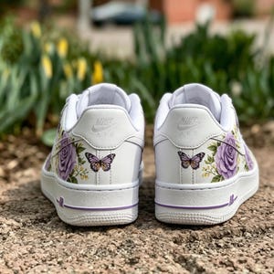Op de afbeelding: Witte sneakers met paarse bloemen- en vlinderontwerpen. De schoenen hebben een low-top design met een witte zool en paarse accenten. Het Nike Air-logo is zichtbaar op de achterkant.