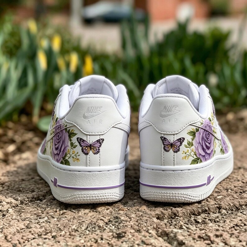 Op de afbeelding: Witte sneakers met paarse bloemen- en vlinderontwerpen. De schoenen hebben een low-top design met een witte zool en paarse accenten. Het Nike Air-logo is zichtbaar op de achterkant.