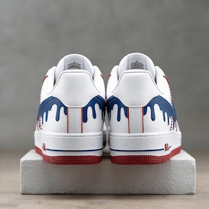 May include: White sneakers with red and blue accents. The design features a white base with blue dripping paint details and red accents on the sole and heel. The sneakers are displayed on a white block.