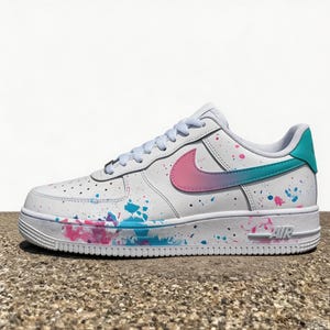 May include: White leather sneaker with a colorful paint splatter design. The Nike swoosh is pink and teal. The heel tab is teal. The sole is white with the word "AIR" in pink.