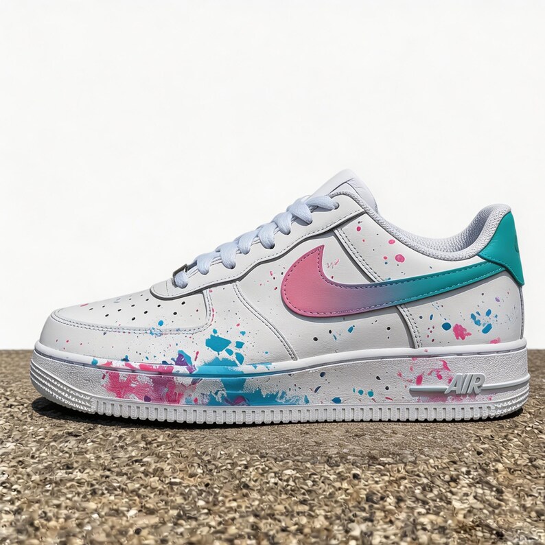 May include: White leather sneaker with a colorful paint splatter design. The Nike swoosh is pink and teal. The heel tab is teal. The sole is white with the word "AIR" in pink.