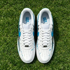 May include: Pair of white sneakers with blue accents and black paint splatter design. The shoes feature blue swooshes and a white lace-up closure. The sneakers are displayed on a bed of green grass.