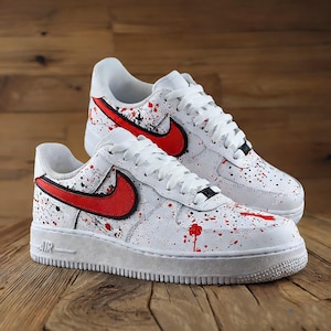 Może przedstawiać: Para białych sneakers&oacute;w z czerwonym i czarnym wzorem rozprysk&oacute;w farby. Posiadają czerwone logo swoosh i białe sznurowadła. Buty mają białą podeszwę i są prezentowane na drewnianej powierzchni.