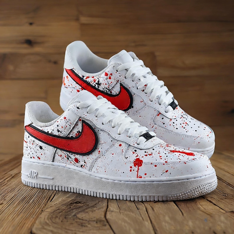 Może przedstawiać: Para białych sneakers&oacute;w z czerwonym i czarnym wzorem rozprysk&oacute;w farby. Posiadają czerwone logo swoosh i białe sznurowadła. Buty mają białą podeszwę i są prezentowane na drewnianej powierzchni.