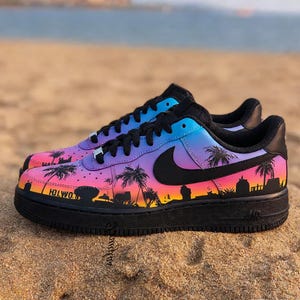 May include: Pair of custom Air Force 1 sneakers with a vibrant sunset design. The shoes feature a black sole, black laces, and a gradient upper with palm trees and the word "HOLWO" in black.