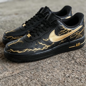Op de afbeelding: Zwarte sneakers met gouden bliksemmotieven en een gouden Nike swoosh. De schoenen hebben zwarte veters en zolen, met het woord "AIR" in gouden letters op de zijkant.