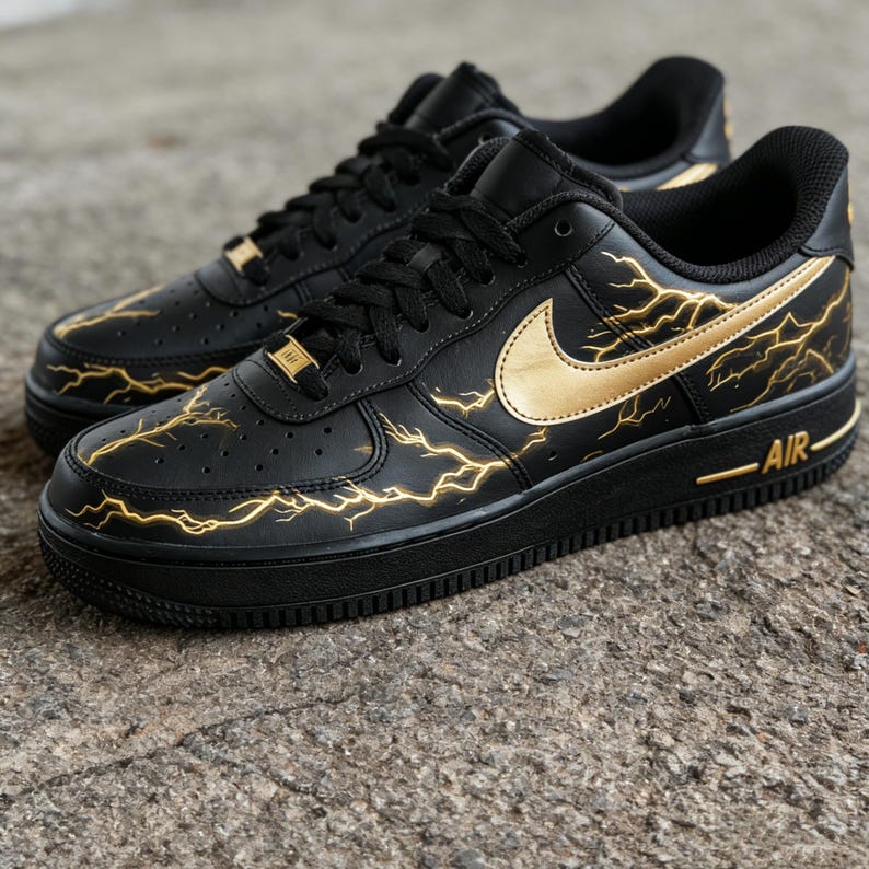 Op de afbeelding: Zwarte sneakers met gouden bliksemmotieven en een gouden Nike swoosh. De schoenen hebben zwarte veters en zolen, met het woord "AIR" in gouden letters op de zijkant.