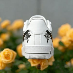 May include: White leather sneaker with black rose graphics on the heel and side. The shoe has a black stripe and a white sole. The background features yellow roses.