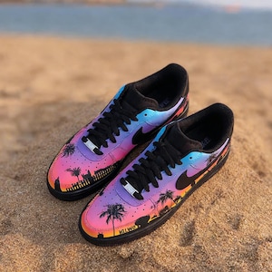May include: Pair of black sneakers with a vibrant sunset-themed design. The shoes feature a gradient of pink, purple, and blue, with a black silhouette of palm trees and buildings. The word "Hollywood" is visible on the side.