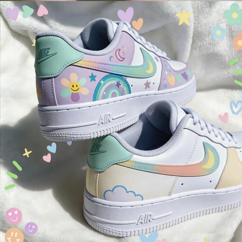 May include: A pair of white sneakers with pastel-colored accents. The shoes feature a light green Nike swoosh and heel tab, with designs including a rainbow, stars, and a smiling flower. The soles are white with "AIR" printed on the side.