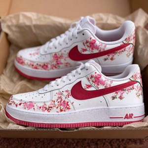 May include: White trainers with a floral pattern. The shoes have a white base, adorned with pink floral designs and a red swoosh. The soles are white with a red trim, and the word "AIR" is visible on the side.