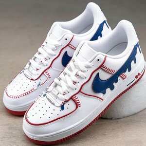 May include: White sneakers with red and blue baseball-themed accents. The shoes feature red stitching, a blue swoosh, and blue paint drip details. The soles are red, and the design incorporates baseball stitching.