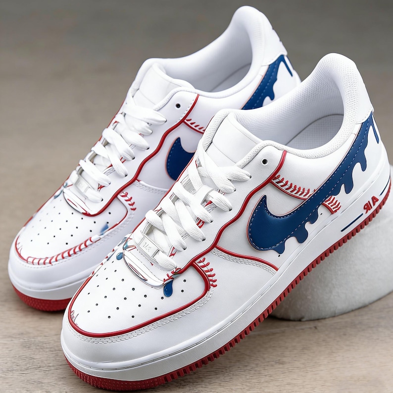 May include: White sneakers with red and blue baseball-themed accents. The shoes feature red stitching, a blue swoosh, and blue paint drip details. The soles are red, and the design incorporates baseball stitching.