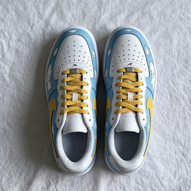 May include: A pair of white, blue, and yellow trainers with a cloud design. The trainers have yellow laces and a yellow swoosh. The shoes are on a white surface.