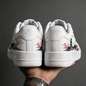 May include: White sneakers with a painted Japanese landscape design. The design features a pagoda, cherry blossoms, and mountains in black and pink. The Nike Air logo is visible on the back of each shoe.