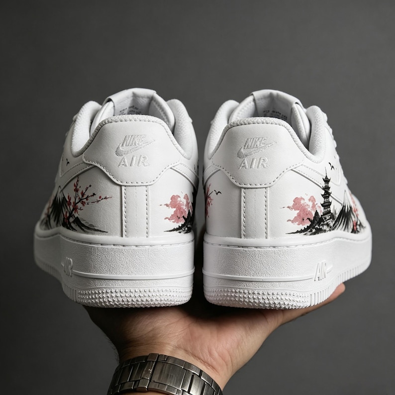 May include: White sneakers with a painted Japanese landscape design. The design features a pagoda, cherry blossoms, and mountains in black and pink. The Nike Air logo is visible on the back of each shoe.