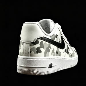 May include: White and gray camouflage pattern sneaker with a black Nike swoosh. The shoe has a white sole with the word "AIR" printed on the side. The sneaker is set against a black background.