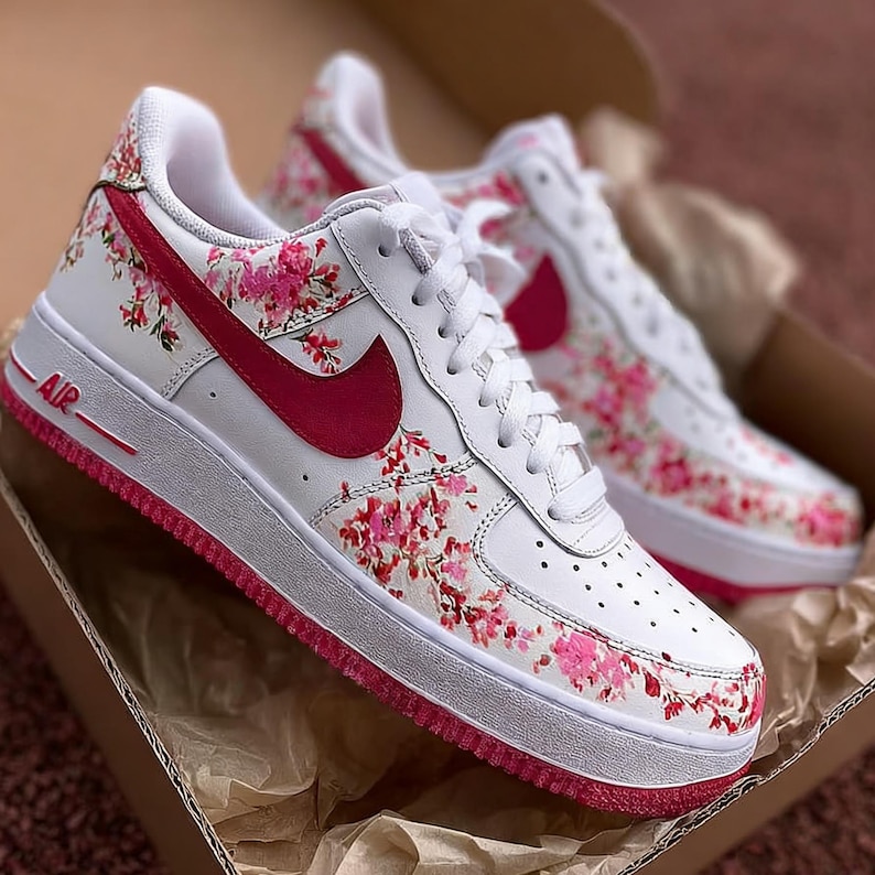 May include: White Air Force 1 trainers featuring a pink floral design. The trainers have a pink Nike swoosh and sole, with the word "AIR" in pink. The shoes are presented in a cardboard box.