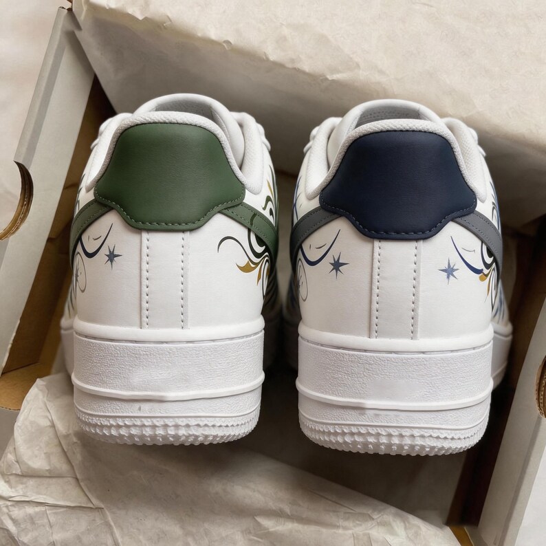 May include: Pair of white sneakers with decorative designs. The heel tabs are green and navy blue. The shoes have gold and blue floral patterns and star accents. The soles are white.
