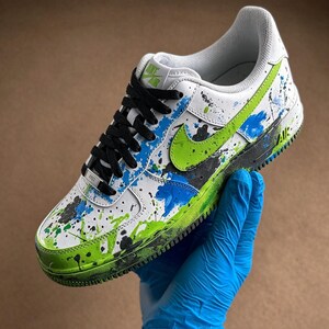 May include: White and green Nike Air Force 1 sneaker with black laces. The shoe features a green swoosh and is decorated with blue, green, and black paint splatters. The sole is green and black.