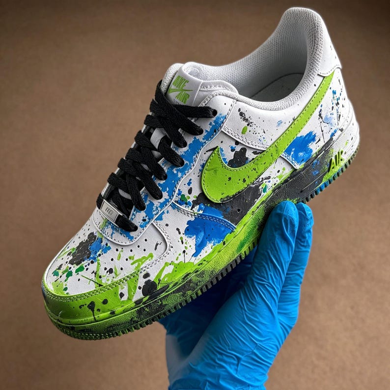 May include: White and green Nike Air Force 1 sneaker with black laces. The shoe features a green swoosh and is decorated with blue, green, and black paint splatters. The sole is green and black.