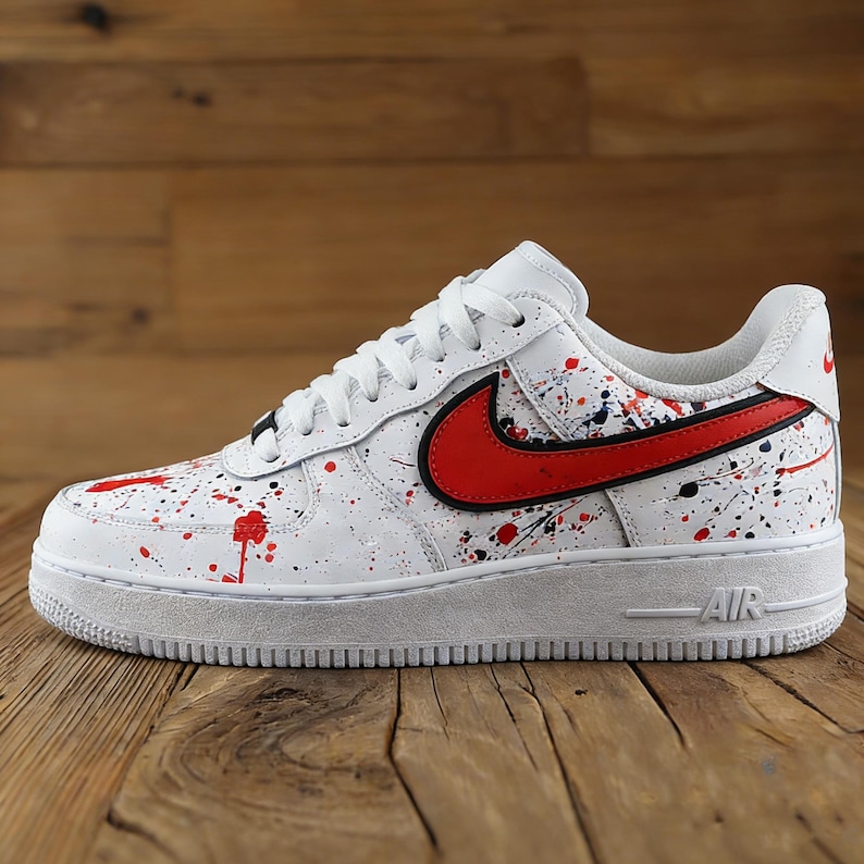 Może przedstawiać: Biały but sportowy z czerwonym i czarnym wzorem rozprysk&oacute;w farby. Posiada czerwone logo swoosh obrysowane na czarno. Podeszwa jest biała ze słowem "AIR" w kolorze czarnym.