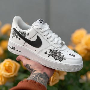 May include: White Air Force 1 sneakers with black rose and leaf designs. The shoe features a black Nike swoosh and the word "AIR" on the side. The background is blurred with yellow flowers.
