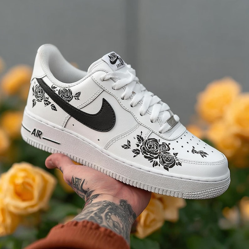 May include: White Air Force 1 sneakers with black rose and leaf designs. The shoe features a black Nike swoosh and the word "AIR" on the side. The background is blurred with yellow flowers.