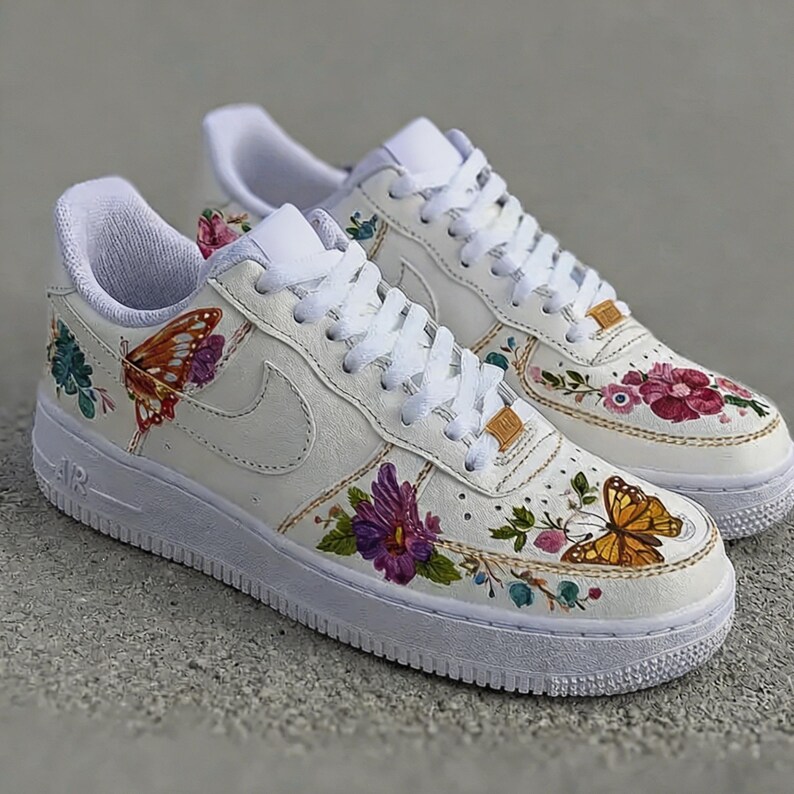 May include: White leather sneakers with floral and butterfly designs. The shoes feature colorful hand-painted flowers and butterflies, with white laces and soles. The iconic Nike swoosh is visible on the side.