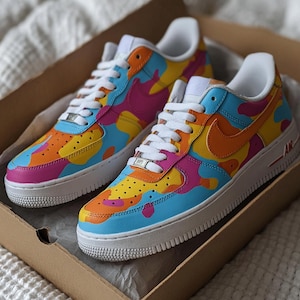May include: A pair of colorful sneakers with a vibrant camouflage pattern in shades of blue, orange, yellow, and pink. The shoes have white laces, soles, and a signature orange swoosh. The sneakers are displayed inside a cardboard box.