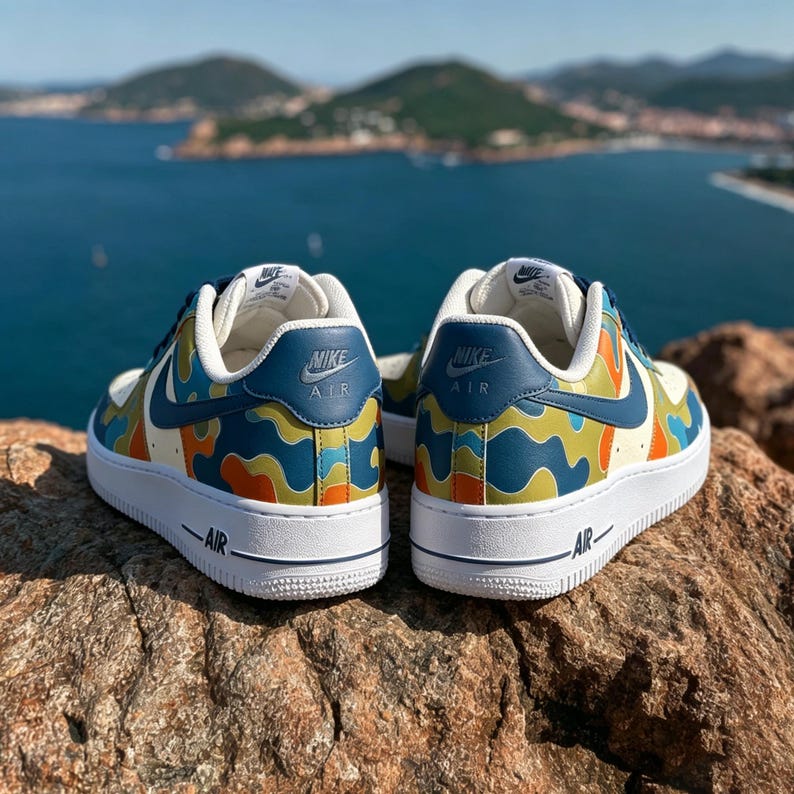 May include: Pair of Nike Air Force 1 sneakers with a colorful camouflage pattern in blue, orange, and green. The shoes have white soles and the Nike Air logo. The background shows a scenic view of the ocean and mountains.