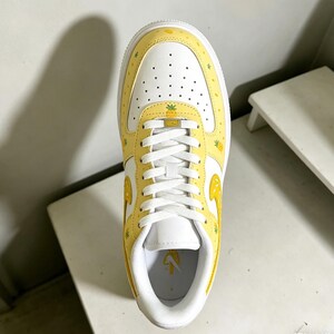 May include: A white and yellow sneaker with a pineapple design. The shoe features a white toe box with small perforations, yellow overlays with pineapple and star patterns, and white laces. The sole is white, and the shoe has a yellow tongue with a pineapple logo.