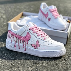 May include: White sneakers with pink accents, including a dripping paint design on the side and a butterfly graphic. The shoes have pink laces and a pink Nike swoosh. The second shoe is visible in the background.