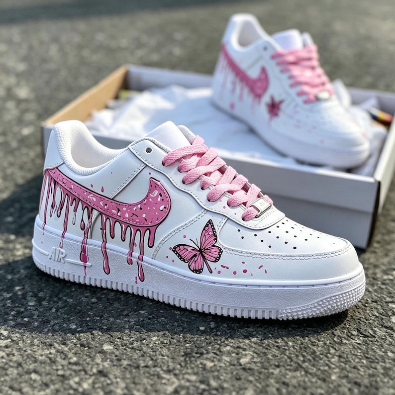 May include: White sneakers with pink accents, including a dripping paint design on the side and a butterfly graphic. The shoes have pink laces and a pink Nike swoosh. The second shoe is visible in the background.